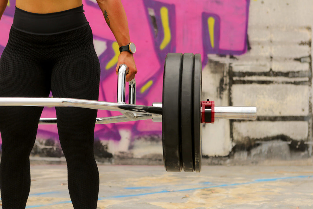 Close-up of a woman lifting a loaded trap bar (hex bar) with weight plates, demonstrating strength training and deadlift exercise using gym equipment.