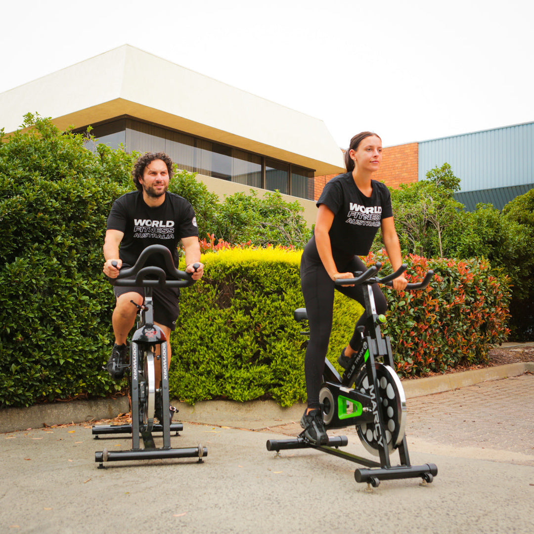 Two people riding stationary spin bikes outdoors in front of a modern building, wearing World Fitness Australia shirts, demonstrating outdoor cardio training and home gym fitness equipment in use.